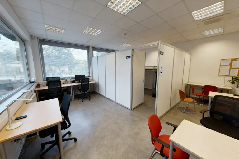 Bureau moderne partagé avec bureaux, cloisons, chaises colorées et grandes fenêtres laissant entrer la lumière naturelle.