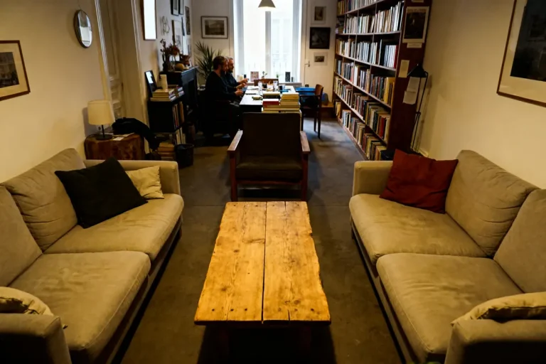 Salon confortable avec deux canapés, table en bois, bibliothèque remplie et deux personnes travaillant au fond.