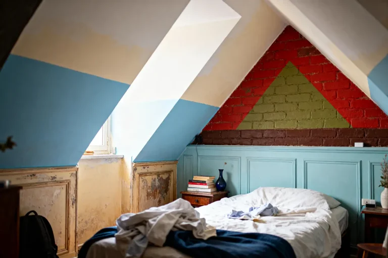Chambre mansardée lumineuse avec lit désordonné, mur en briques coloré, table de chevet et livres empilés.
