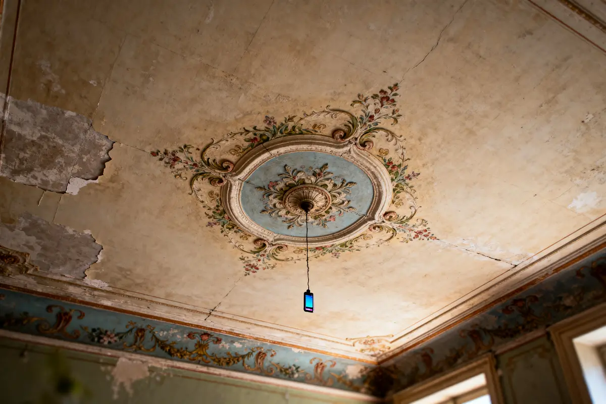 Plafond ancien avec moulures décoratives et téléphone suspendu, peinture dégradée, lumière naturelle par fenêtres.