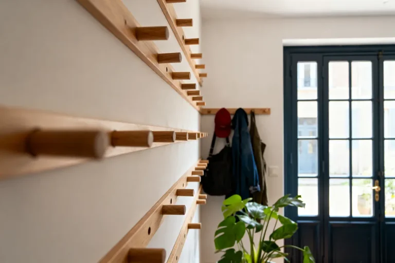 Mur de rangement en bois avec crochets, vestes suspendues, plante verte et porte vitrée en arrière-plan.