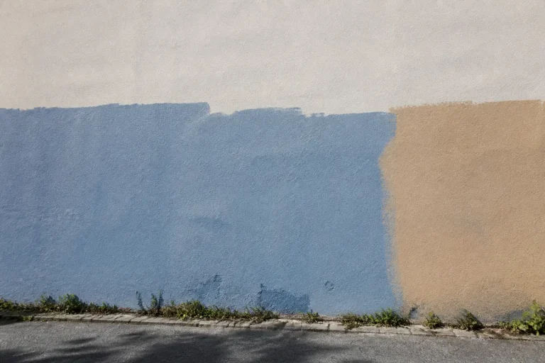Mur partiellement peint en bleu et beige, avec des mauvaises herbes en bas sur un trottoir.