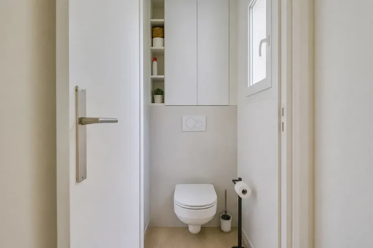 Toilettes modernes avec cuvette suspendue, rangement mural, papier toilette, accessoires minimalistes, fenêtre apportant lumière naturelle.