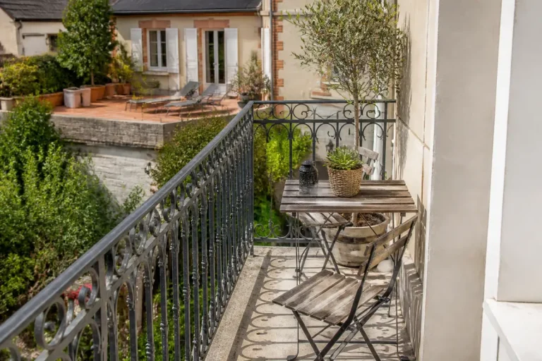 Balcon avec table et chaise en bois, plantes en pot, vue sur terrasse ensoleillée et jardin verdoyant.