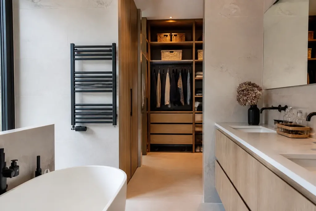 Salle de bain moderne avec baignoire, meuble en bois clair, sèche-serviettes et placard ouvert.