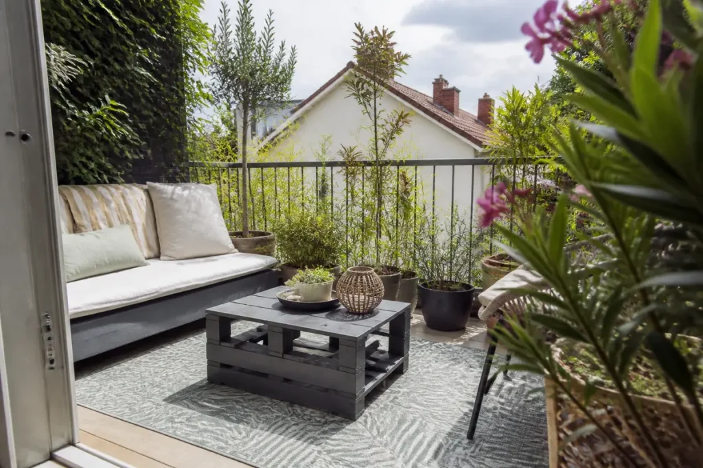 Balcon confortable avec canapé, table basse en bois, tapis, plantes en pots et vue sur maison en arrière-plan.