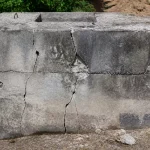 Mur en béton fissuré, entouré de verdure, avec des motifs d'usure visible sur sa surface grise.