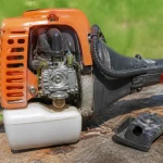 Moteur de débroussailleuse orange posé sur une souche de bois avec fond herbeux flou.