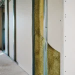 Structure de cloison en construction avec isolant visible, panneau en placoplâtre, ossature métallique, fenêtre en arrière-plan.