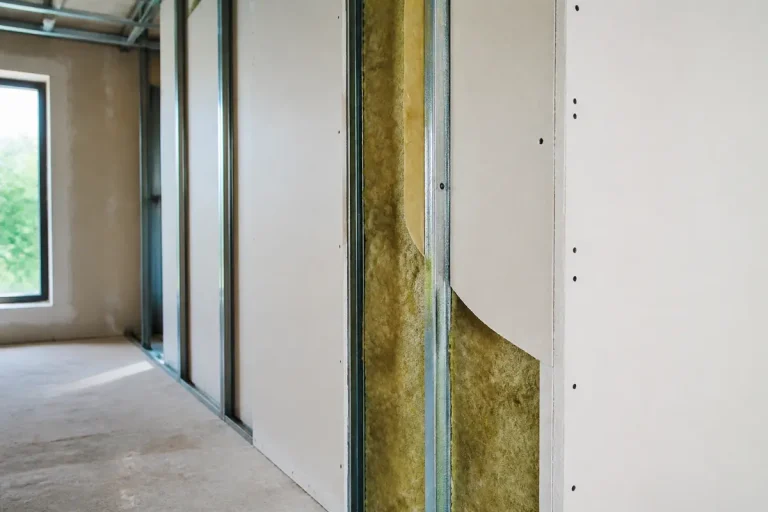 Structure de cloison en construction avec isolant visible, panneau en placoplâtre, ossature métallique, fenêtre en arrière-plan.