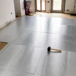Installation de panneaux isolants au sol dans une pièce avec marteau et parquet visible à gauche.