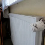 Radiateur blanc sous fenêtre, avec un thermostat réglable sur le côté droit, près d'un bureau en bois.