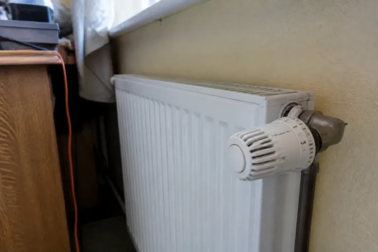 Radiateur blanc sous fenêtre, avec un thermostat réglable sur le côté droit, près d'un bureau en bois.