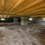 Espace sous une maison avec vide sanitaire, sol en terre battue et piliers béton.