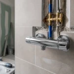 Installation d'un mitigeur mural dans salle de bains rénovée avec murs en carrelage gris.
